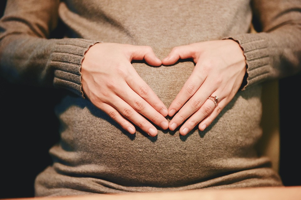 Pregnant Woman In Brown Sweater Holding Hands With Fingers In a Heart Shape Over Abdomen